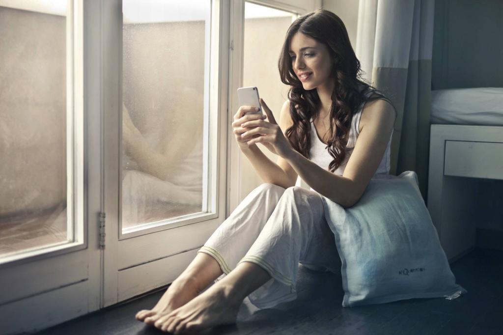 Frau in weißem Sommerkleid sitzt am Fester ihrer Wohnung und schaut auf ihrem Handy und lacht, weil sie bei Frauen anschreiben Vorlagen eine schöne Nachricht bekommen hat. 